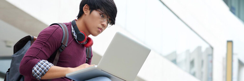 Student with headphones and backpack using laptop outdoors